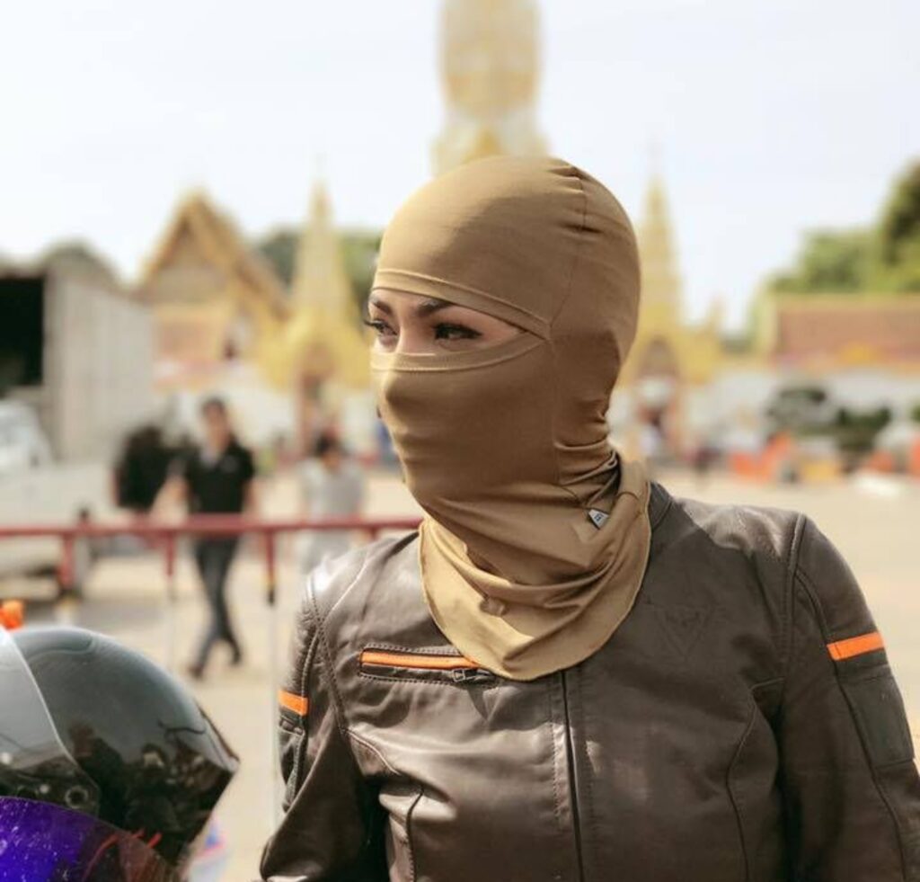A person wearing a tan balaclava that covers their head and neck, with only the eyes visible through a slit. They are dressed in a dark riding jacket with orange reflective accents, standing near a blurred outdoor area with people and structures in the background.