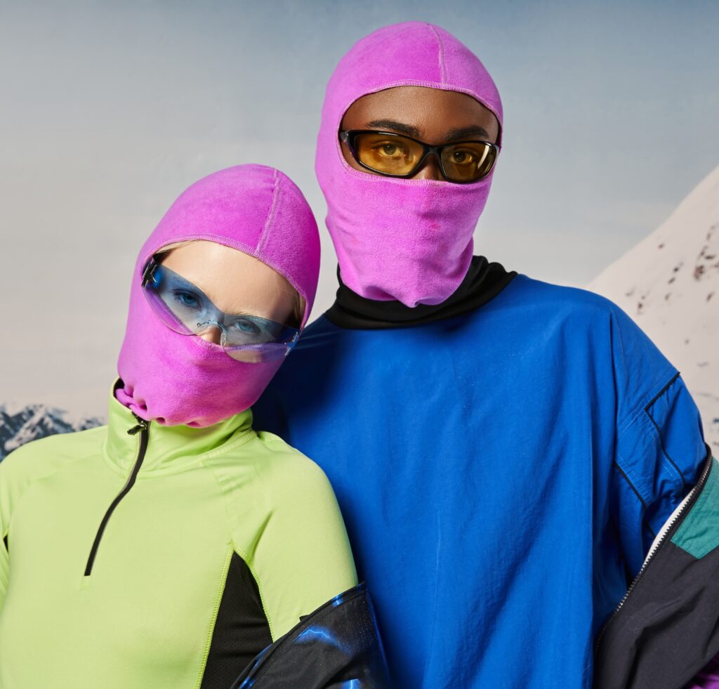 "Two models wearing bright balaclavas: one in a neon pink balaclava with matching pink hood, yellow-tinted sunglasses, and a lime-green zip-up top; the other in a pink balaclava with lavender lenses, a blue oversized top, and protective eyewear. Snowy mountainous backdrop."