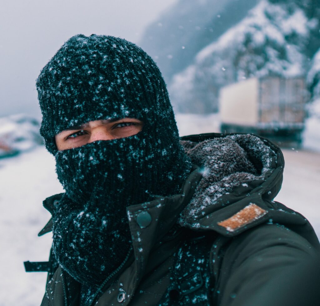 A person wearing a dark balaclava and winter jacket stands outdoors in a snowy, windy environment, with only their eyes visible. Snow covers the balaclava and shoulders, and a snow-covered road and trucks are blurred in the background.