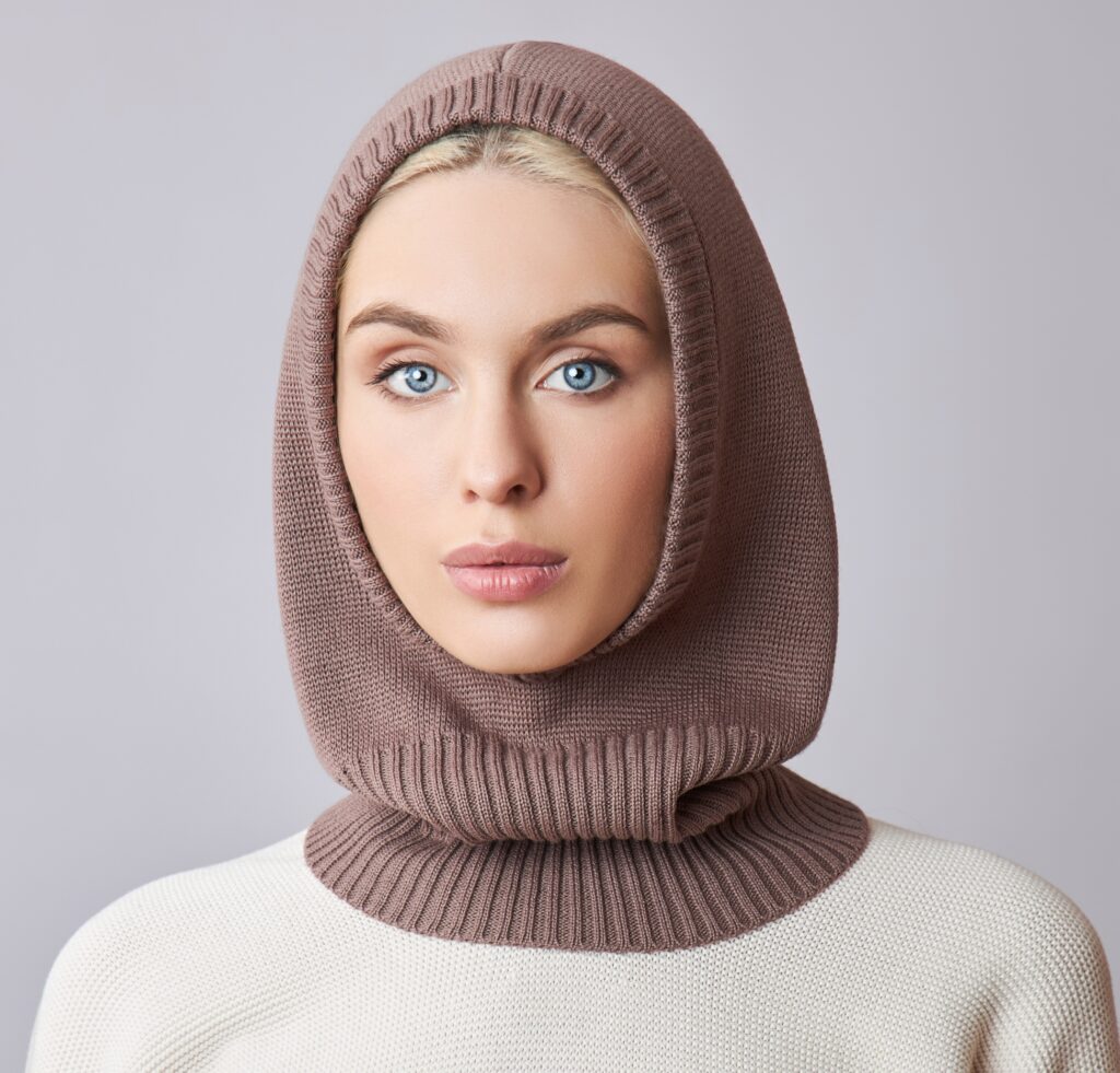 A close-up portrait of a woman wearing a soft, dusty pink open-face wrap balaclava. The knit hood frames her face with a high ribbed edge around the chin and neck, and she wears a light beige sweater. Her blue eyes and neutral makeup are visible, with the balaclava framing her features.