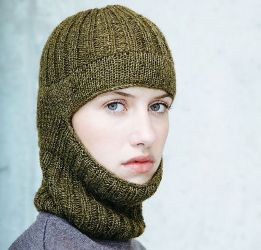 A young person wearing a green knitted balaclava that covers the head, neck, and face except for the eyes, nose, and mouth. The neutral background is blurred.