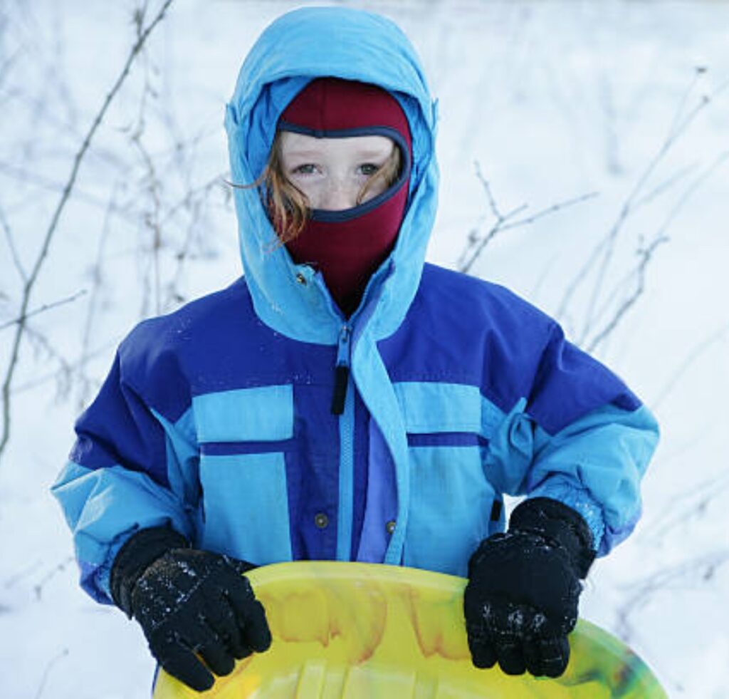 A child dressed in a blue winter jacket, red balaclava covering their face except for the eyes, standing in a snowy landscape and holding a yellow sled.