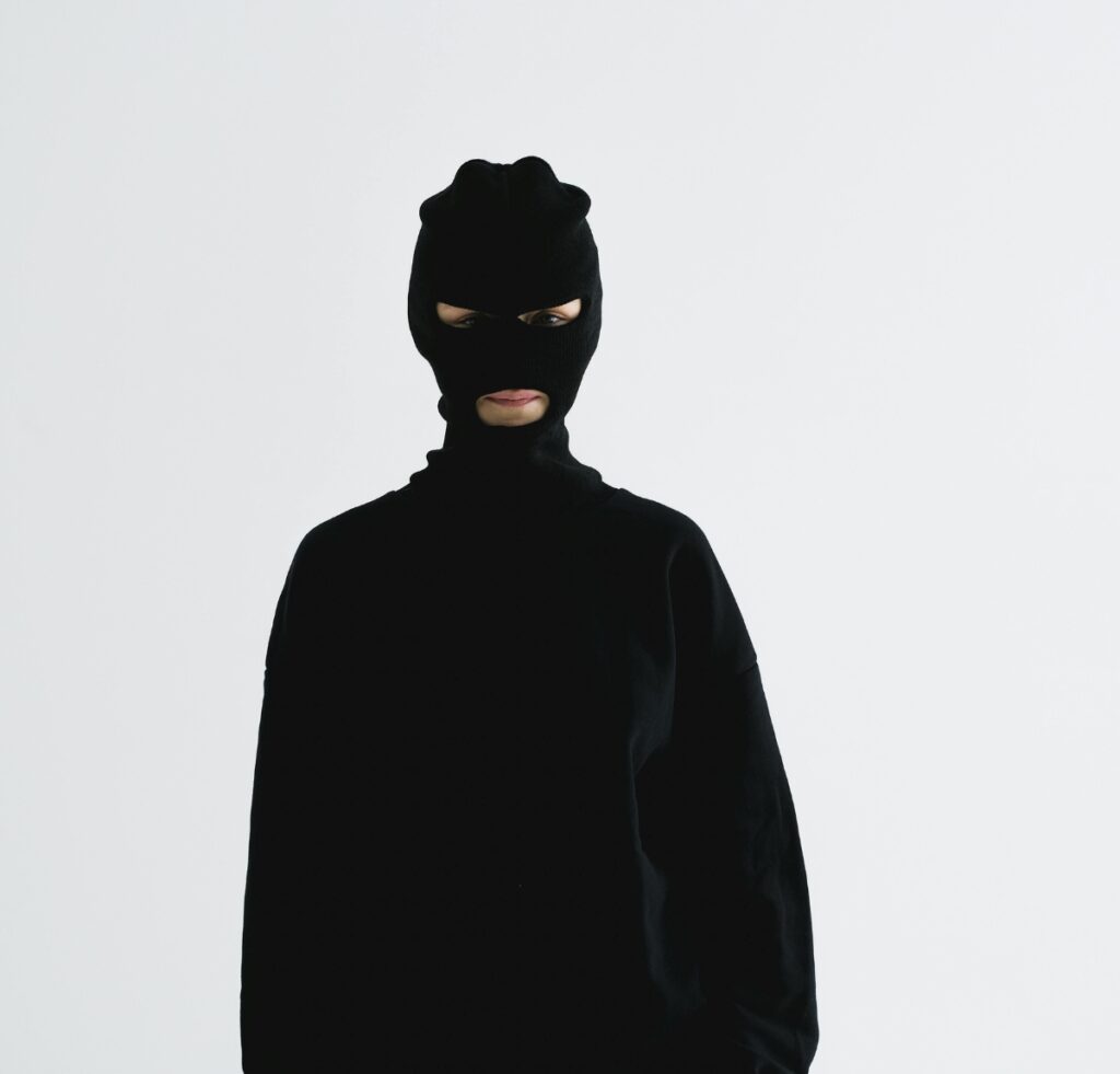 A person wearing a black balaclava and black long-sleeve top, with their face largely obscured except for the eyes and mouth, standing against a light background.