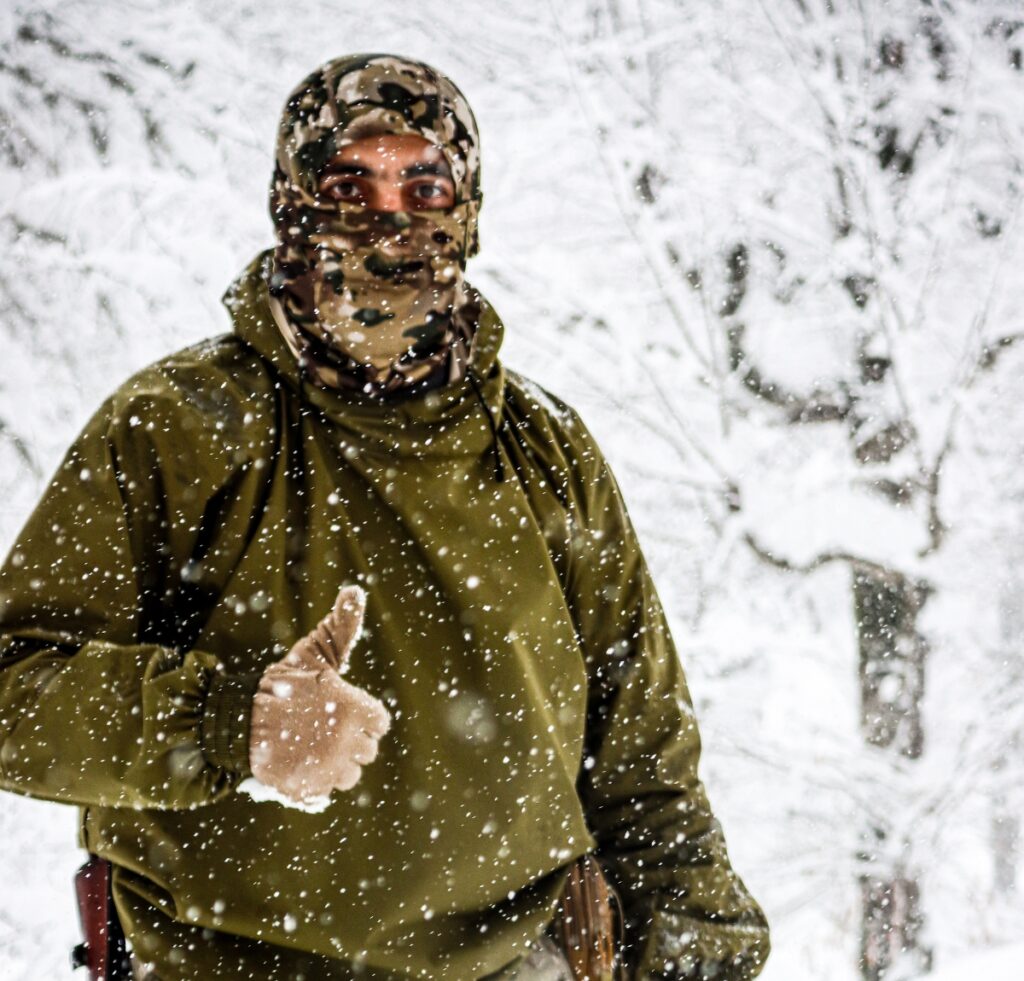 A person in a camouflage balaclava and olive jacket stands in a snowy forest, giving a thumbs-up as snow falls around them.
