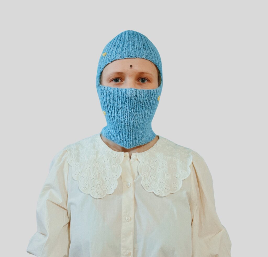 A person wearing a blue knitted balaclava that covers the head and neck, leaving only the eyes exposed, over a cream-colored blouse with a decorative white collar.