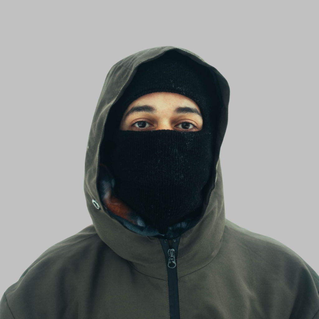 A person wearing a dark hooded jacket with the hood up and a black balaclava covering the lower face, leaving the eyes visible.