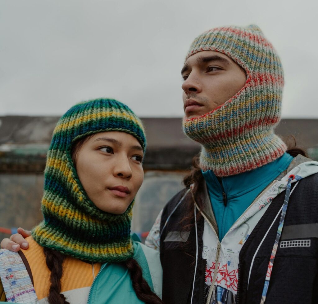 A close-up photo of two people wearing knitted balaclavas. The person on the left is wearing a green, yellow, and blue striped balaclava that covers the head and neck but leaves the face exposed. The person on the right is wearing a multicolored knit balaclava with the face opening around the mouth and nose; they also wear a blue jacket with layered outerwear. The background is urban and slightly blurred.