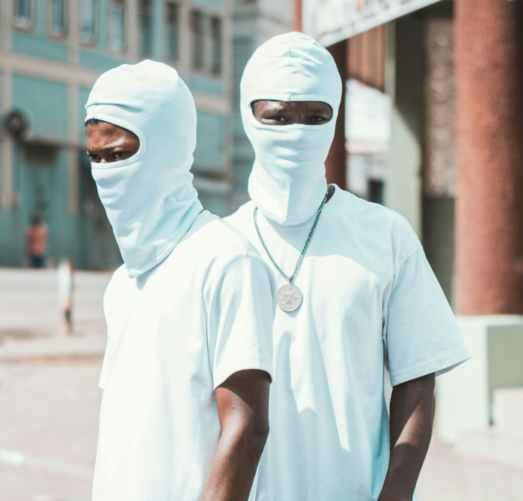 Two people wearing light blue balaclavas and matching t-shirts, standing on a sunlit urban street; one person faces the camera while the other looks away, both wearing balaclavas that cover their heads and necks with only the eyes visible.
