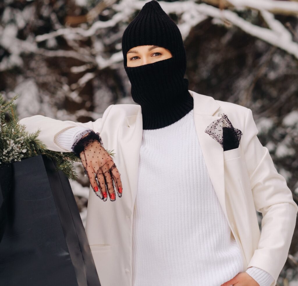 A stylish person wearing a black balaclava, white knit sweater, and an off-white blazer with a dotted mesh pocket square. They lean on a large black bag outdoors in a snowy setting, with evergreen branches nearby.
