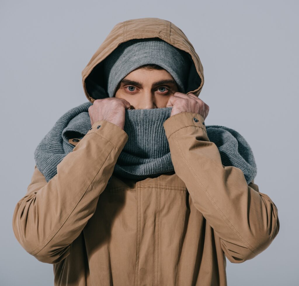 A person wearing a tan jacket and gray knit cap, pulling a gray scarf up to cover their lower face like a balaclava.