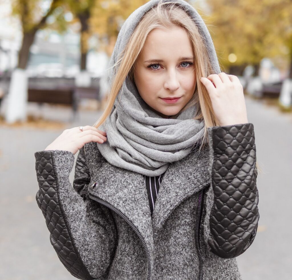 A young woman wearing a light gray scarf wrapped around her neck and a gray textured coat with quilted black sleeves, standing outdoors on a fall day.