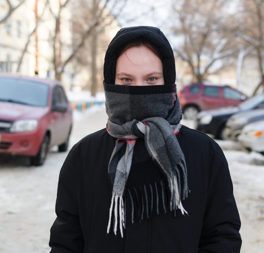 A person outdoors in a winter scene wearing a black hood and a gray-black scarf wrapped around the lower half of the face, with fringed ends visible. Snowy street and parked cars in the background.