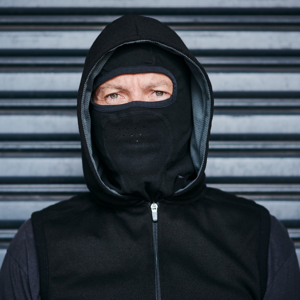 A person wearing a black balaclava with only the eyes visible, zipped hoodie, standing in front of a striped metal shutter