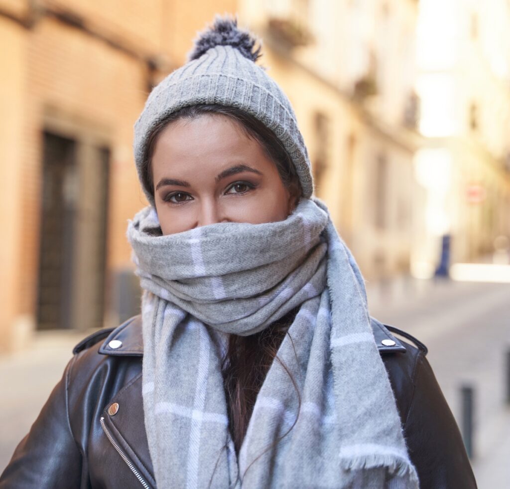A stylish young woman outdoors in a light gray beanie and matching scarf wrapped around her neck and lower face, wearing a dark leather jacket. The background shows a sunlit street with warm-toned buildings.