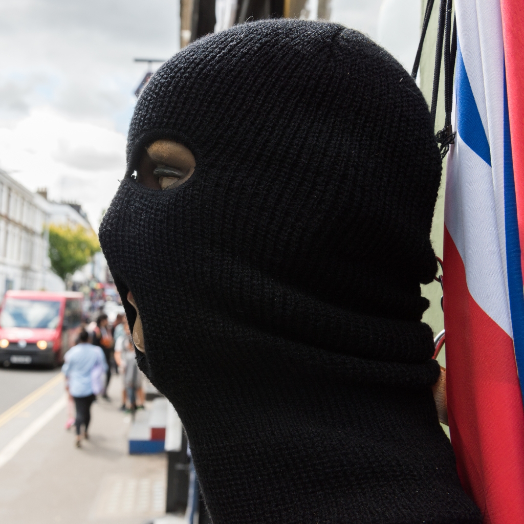 A person wearing a black knit balaclava covering most of their head and face, with only one eye visible, standing on a busy city sidewalk near colorful flags.
