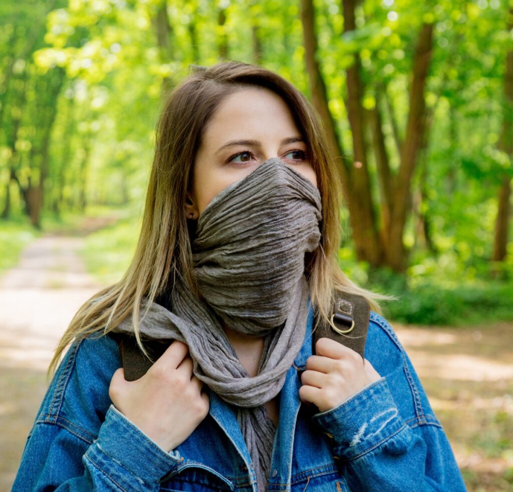 A woman standing outdoors in a green forest, wearing a gray scarf wrapped around her mouth and nose, and a blue denim jacket. She holds the scarf and backpack straps, and the path behind her is dappled with sunlight.