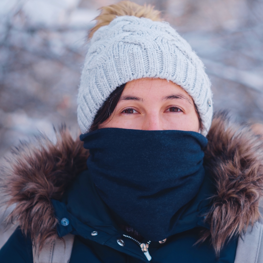 “A person dressed for cold weather wearing a light gray knit beanie and a dark Scarf balaclava that covers the lower face, with a fur-trimmed hood and winter jacket outdoors in a snowy setting.”