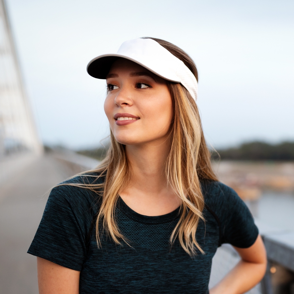 A woman wearing a white visor cap and a dark athletic t-shirt stands on a waterfront boardwalk, looking off to the side with a subtle smile.
