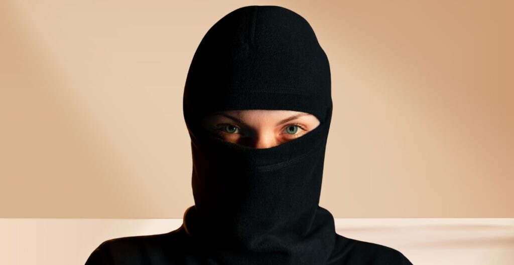 A person wearing a black balaclava, with only their eyes visible, posing against a light tan background.