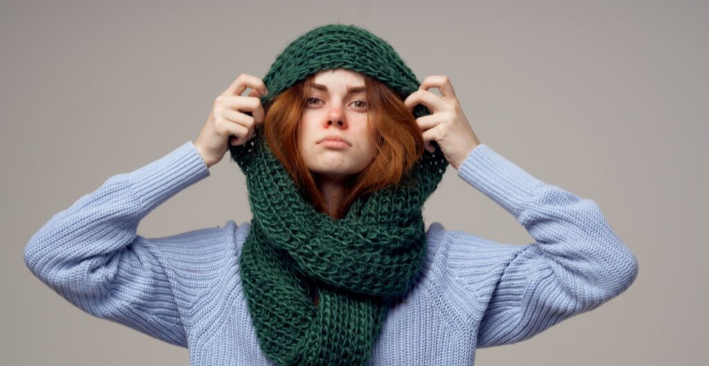 "Person wearing a green knitted balaclava-style scarf over a light blue sweater, adjusting the hood with their hands