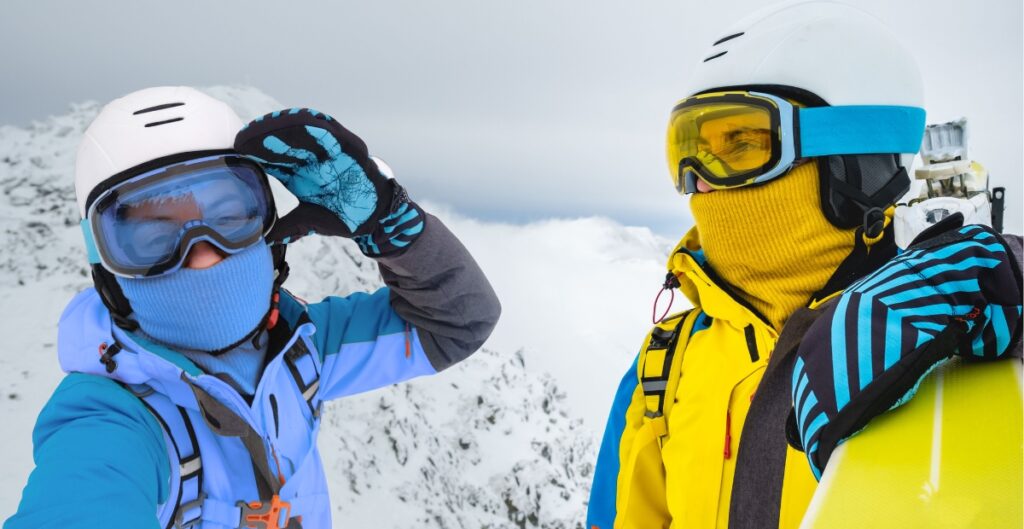Two skiers in balaclavas and winter gear stand on a snowy slope, one adjusting their goggles.