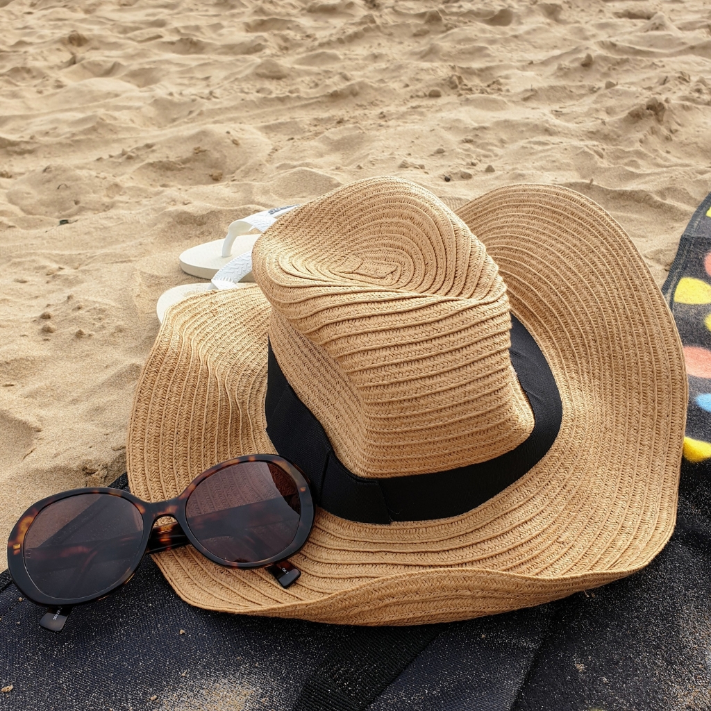 A sandy beach scene featuring a straw sun hat with a black band, a pair of dark round sunglasses resting on the brim, and a glimpse of flip-flops in the background on light tan sand.

