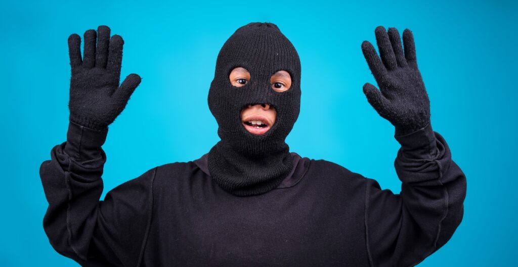 “A person wearing a black ski mask and black clothing stands against a bright blue background, with both hands raised.”