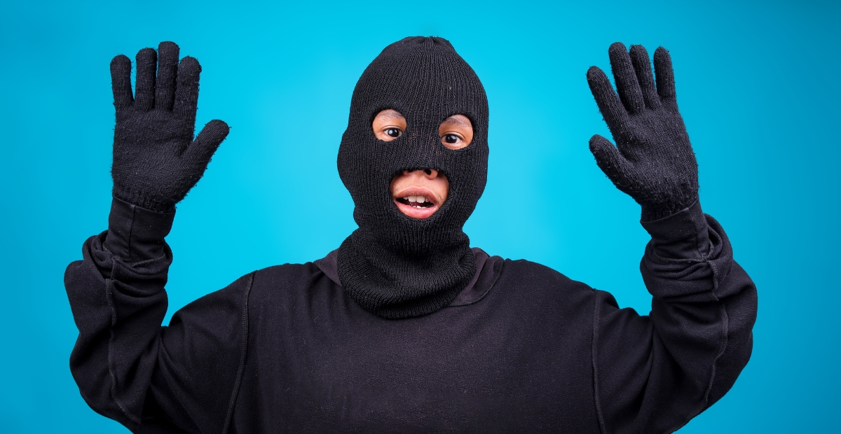 “A person wearing a black ski mask and black clothing stands against a bright blue background, with both hands raised.”