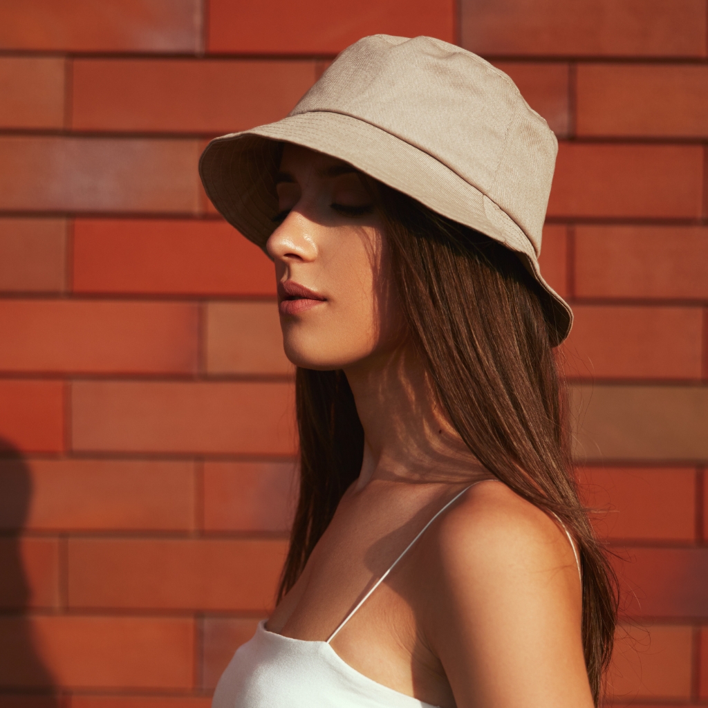 Eine junge Frau mit einem hellbeigen Bucket Hat und einem weißen Top mit Spaghettiträgern steht vor einer roten Backsteinmauer, die Augen geschlossen, das Profil nach links gewandt.
