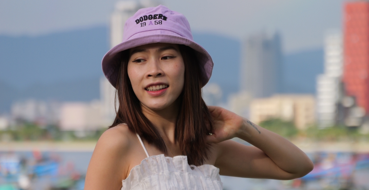 Eine junge Frau mit einem hellvioletten Bucket Hat und einem weißen, ärmellosen Oberteil steht im Freien, im Hintergrund sind verschwommen die Skyline einer Stadt und Hügel zu sehen.