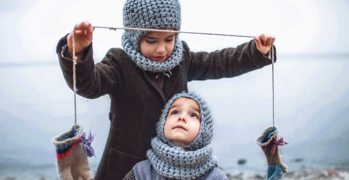 Eine Person steht hinter einem Kind und hält eine Schnur mit zwei gestrickten Handschuhen daran; beide Kinder tragen kuschelige graue Sturmhauben und gestrickte Schals.