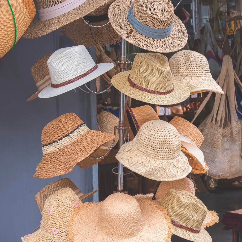 A display of various woven beach hats hanging on a stand, including wide-brimmed straw hats in natural tones and a white fedora with a brown band.

