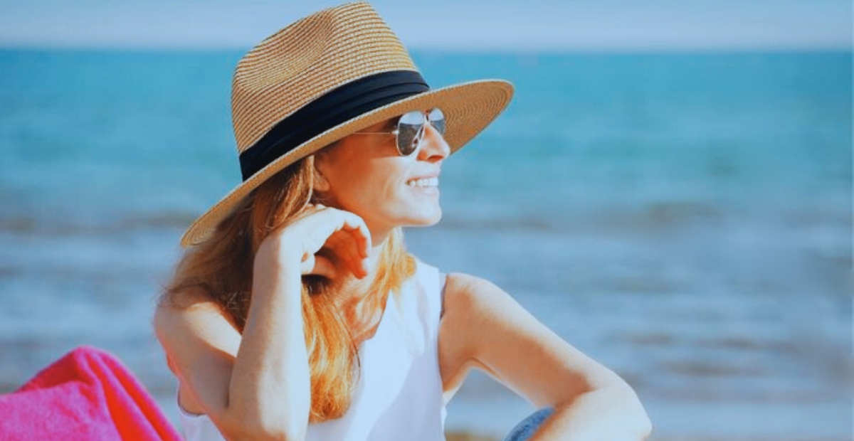 „Frau am Strand mit einem breitkrempigen, hellbraunen Fedora-Hut mit schwarzem Band, Sonnenbrille und einem weißen, ärmellosen Oberteil; im Hintergrund das Meer.“
