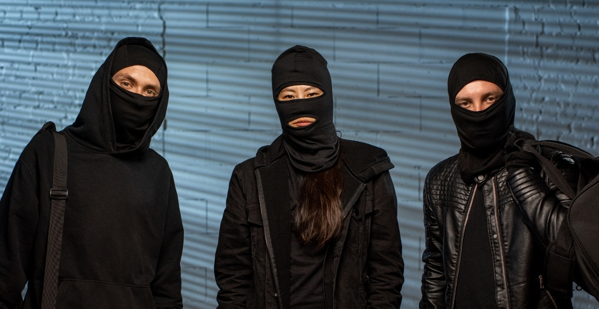 Three people wearing black balaclavas and dark clothing standing against a blue-gray brick wall.