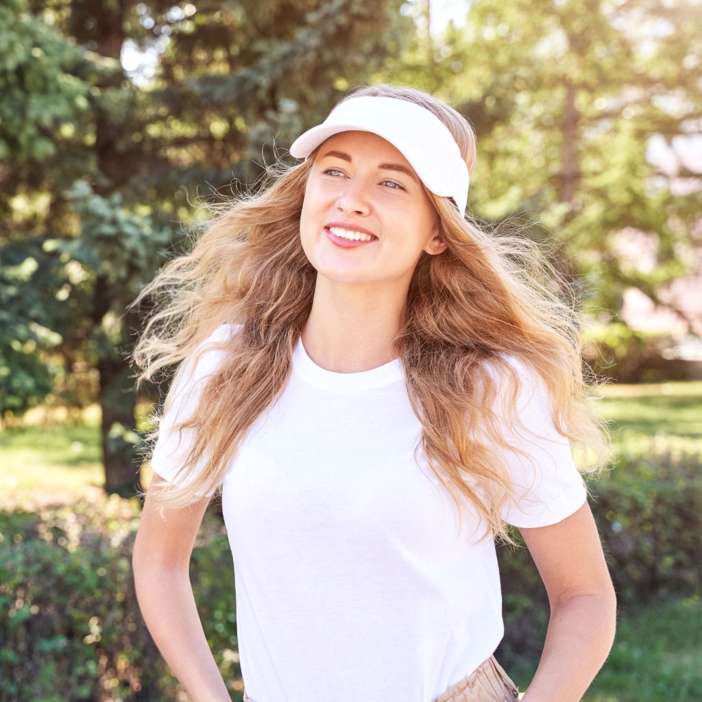 A smiling young woman wearing a white cotton visor, white t-shirt, and light-colored shorts stands outdoors in a sunny park with green trees in the background.