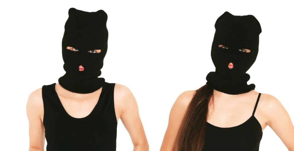 Two people wearing black balaclavas that cover their heads and most of their faces, with only their eyes and mouths visible. Both are dressed in black tops; the person on the left has a sleeveless shirt, the person on the right has a black spaghetti-strap top.