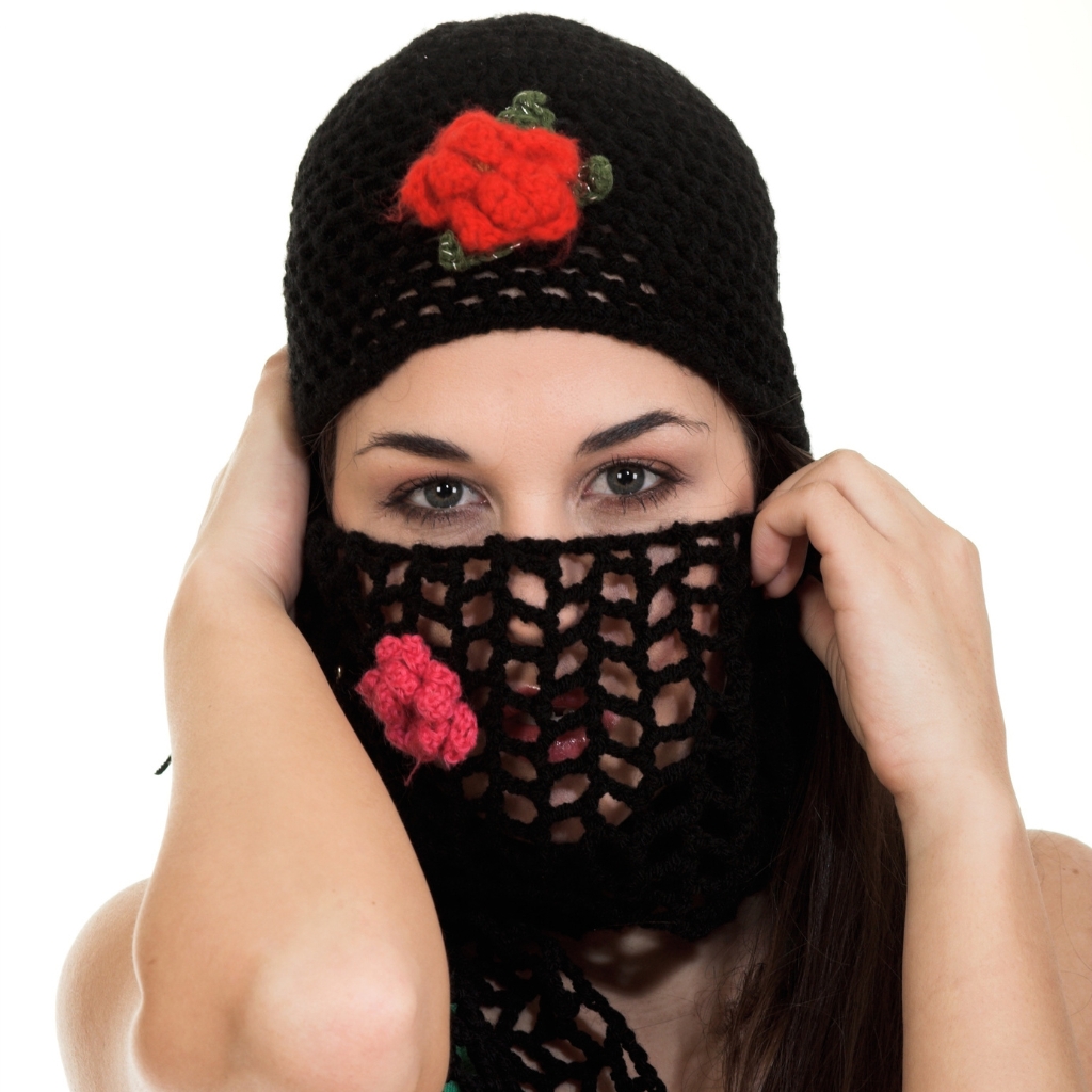 A woman wearing a black knitted balaclava with a lacy, open-weave section over the lower face; a bright red knitted flower decorates the hat, and the woman’s eyes are visible through the mesh.