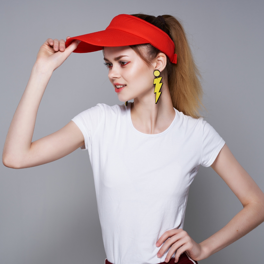 A stylish woman wearing a red visor, white tee, and yellow zigzag ear earrings, posing with one hand on her hip against a neutral backdrop