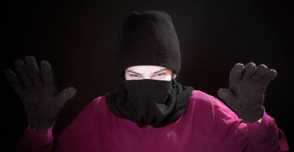 A person wearing a black balaclava, purple shirt, and black knit gloves, with hands raised against a dark background