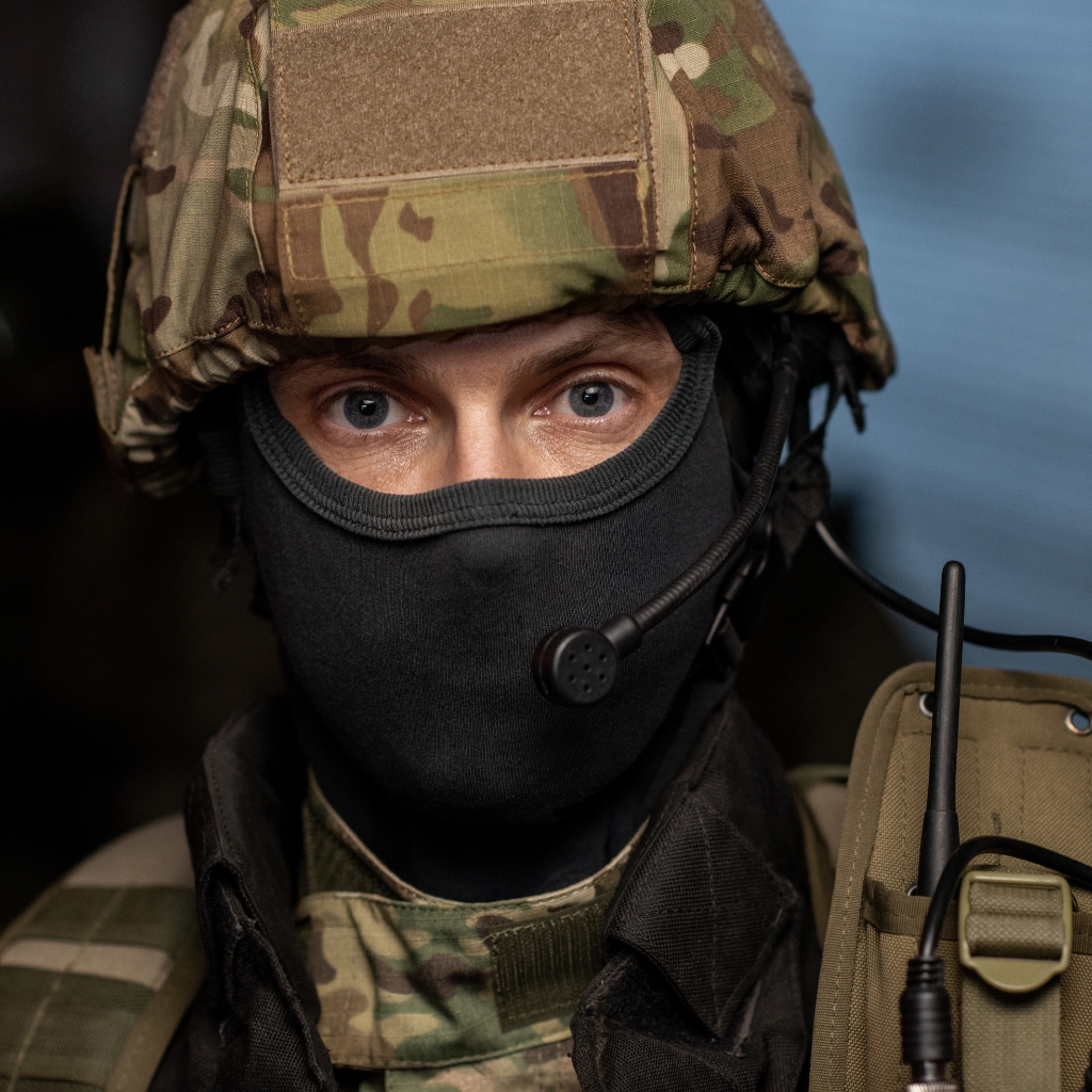 A close-up of a soldier in full camouflage gear, wearing a helmet and a black balaclava with a headset microphone.