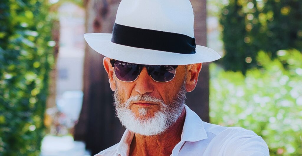 Nahaufnahme von einem älteren Mann, der einen weißen Panamahut mit einem schwarzen Band, eine Sonnenbrille und und einem weißen Hemd, im Freien mit Grünpflanzen im Hintergrund .