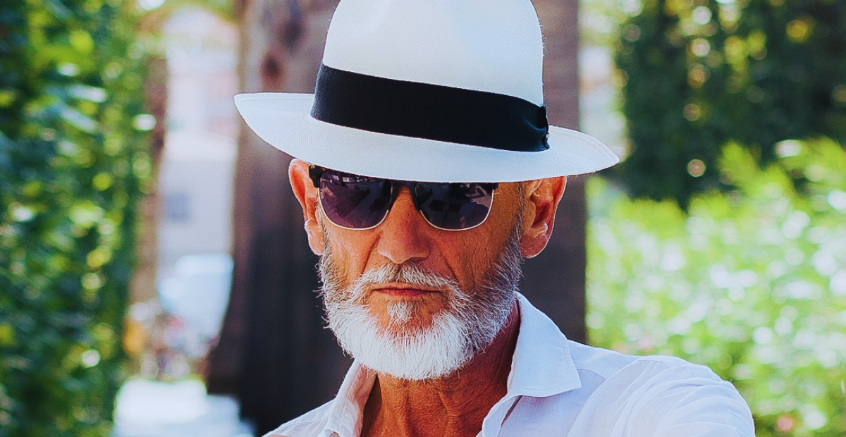 Gros plan d'un homme âgé portant un chapeau Panama blanc avec un ruban noir, des lunettes de soleil et une chemise blanche, à l'extérieur avec de la verdure en arrière-plan.