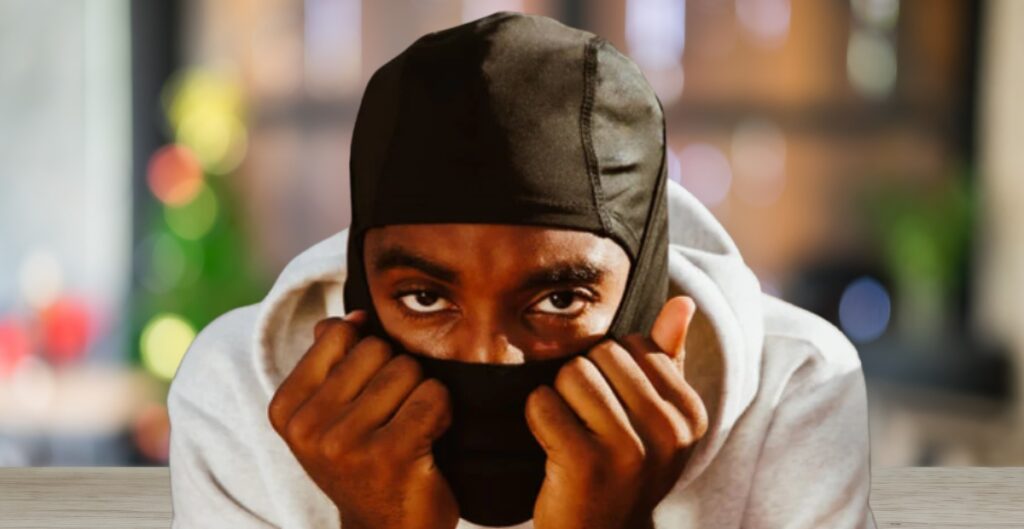 A person wearing a black balaclava and a light gray hoodie sits at a wooden table, hands near their face, looking directly at the camera with intense eyes