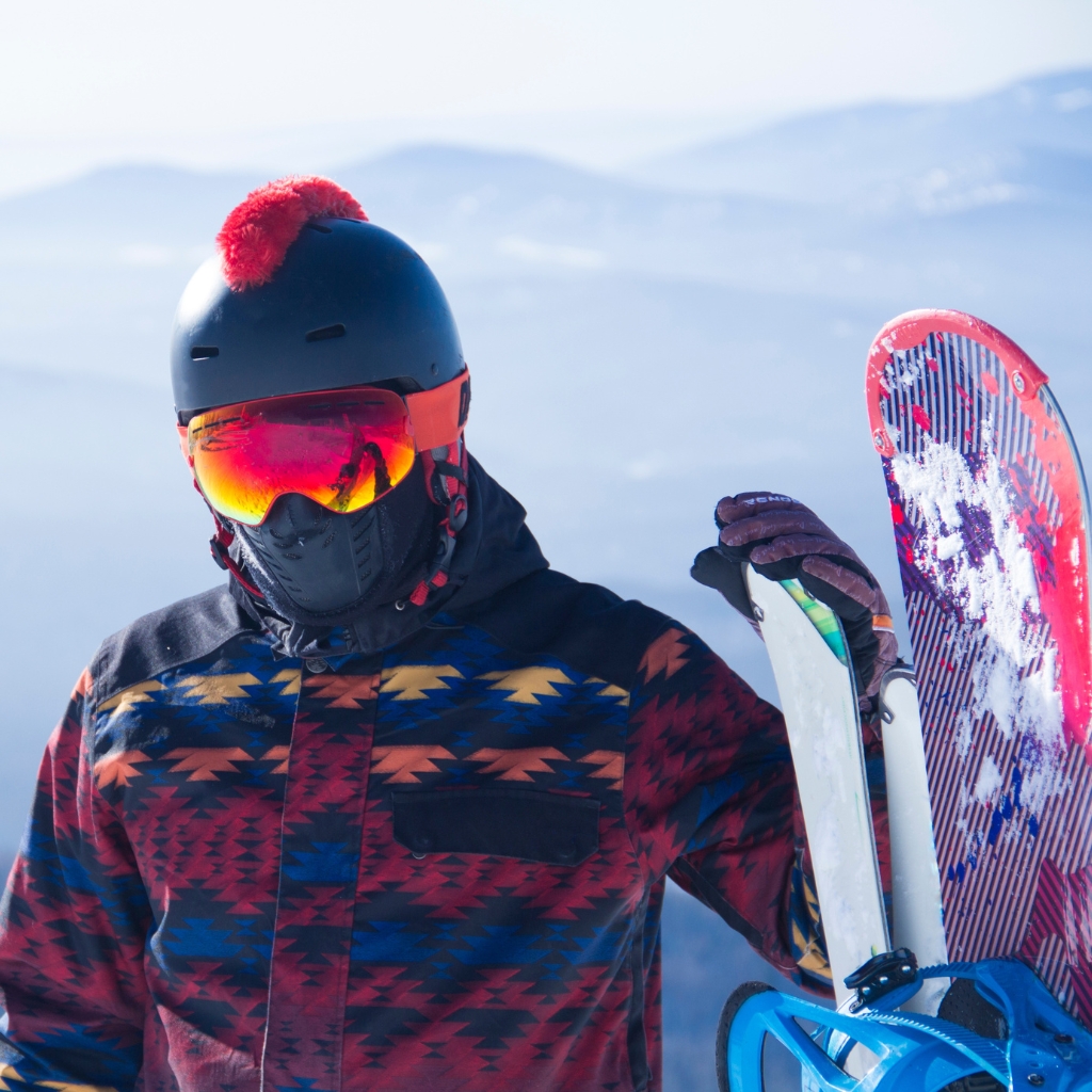A skier wearing a dark helmet with a red pom-pom, bright orange-tinted goggles, and a patterned jacket, holding a snowboard with bindings in a snowy mountain setting.
