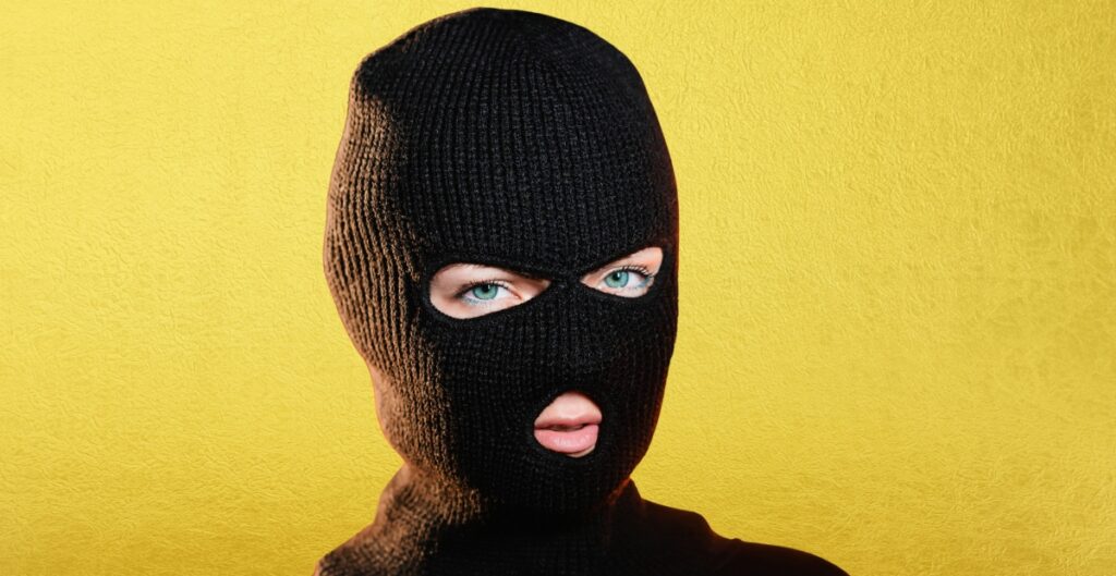 Person wearing a black balaclava with only the eyes and mouth visible, against a bright yellow background.