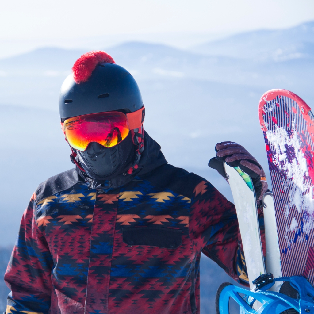 A snowboarder wearing a black helmet with a red pom-pom, reflective orange goggles, a black face mask, and a colorful geometric-patterned jacket stands in a snowy alpine setting holding a pair of skis and a snowboard upright beside them.
