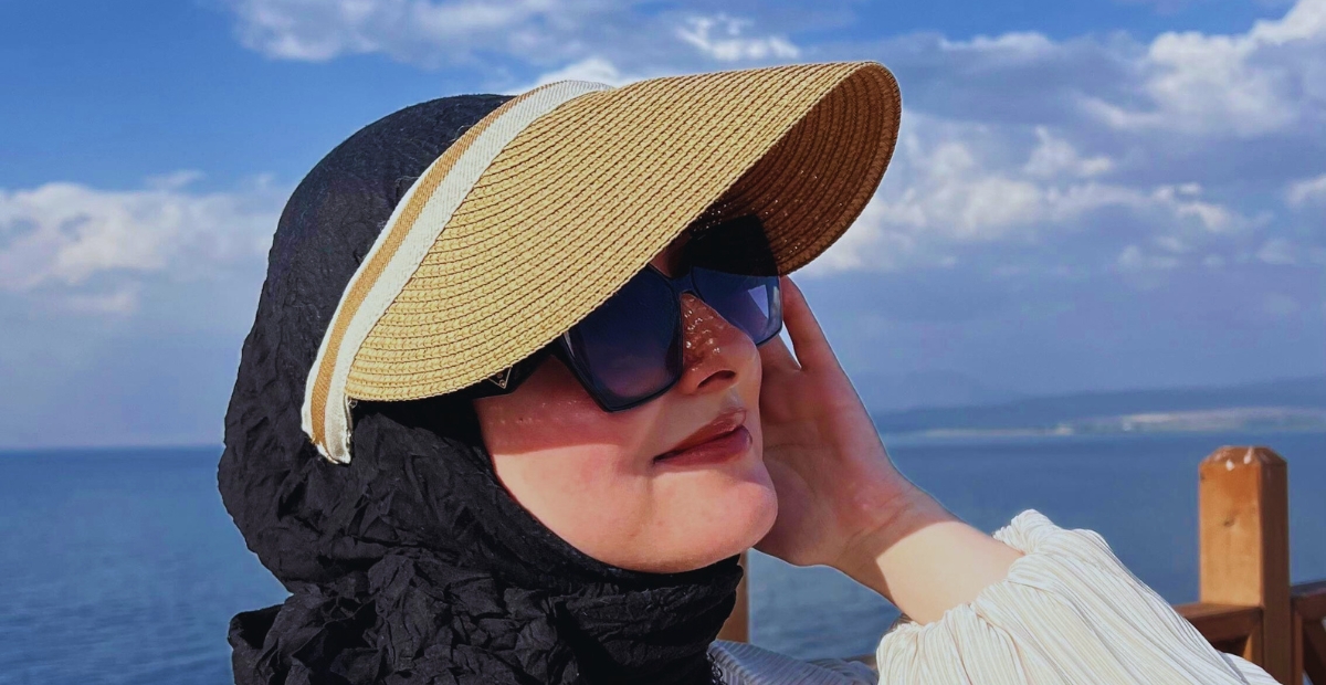 Une femme portant un grand chapeau à visière en paille, des lunettes de soleil foncées et un chemisier de couleur claire, debout près d'un plan d'eau avec une balustrade en bois en arrière-plan. Elle sourit légèrement et touche le côté de son visage d'une main. Un foulard noir couvre ses cheveux.