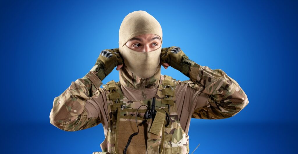 A soldier wearing camouflage uniform and a tan balaclava covers most of his face, with only the eyes visible. He is adjusting the balaclava with both hands, standing against a solid blue background.