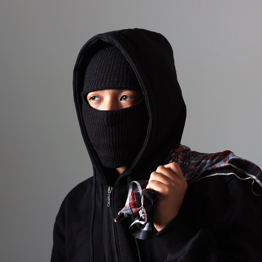 A person wearing a black balaclava, hoodie, and gloves. Only their eyes are visible through the knit face covering, and they’re holding a plaid shirt over their shoulder.
