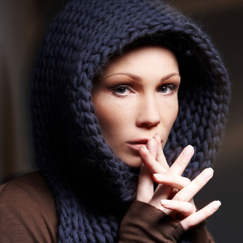 Nahaufnahme einer Frau, die eine dicke, marineblaue Häkel-Sturmhaube trägt, die ihr Gesicht umrahmt; sie ist leicht geschminkt und hat die Hände vor dem Mund gefaltet.
